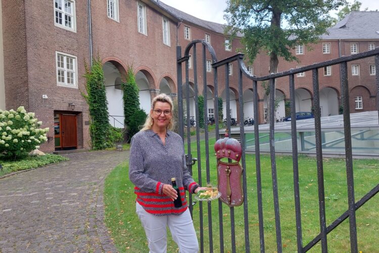 Gästeführerin Birgit van den Boom steht vor dem schmiedeeisernen Tor mit einem roten Wappen, im Hintergrund ein Teil der Schwanenburg in Backstein und ein gepflasterter Weg