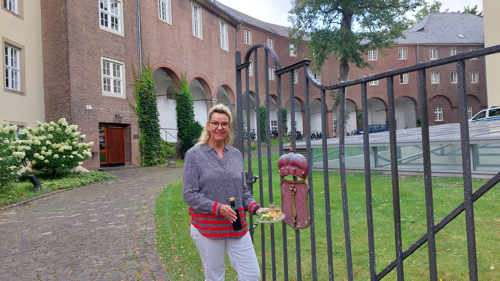 Gästeführerin Birgit van den Boom steht vor dem schmiedeeisernen Tor mit einem roten Wappen, im Hintergrund ein Teil der Schwanenburg in Backstein und ein gepflasterter Weg