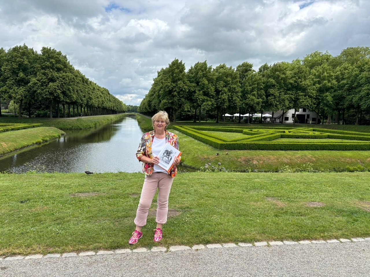 Gästeführerin Elisabeth Thönnissen steht auf einer Wiese vor dem Moritzkanal in den historischen Gartenanlagen mit Baumreihen und Hecken