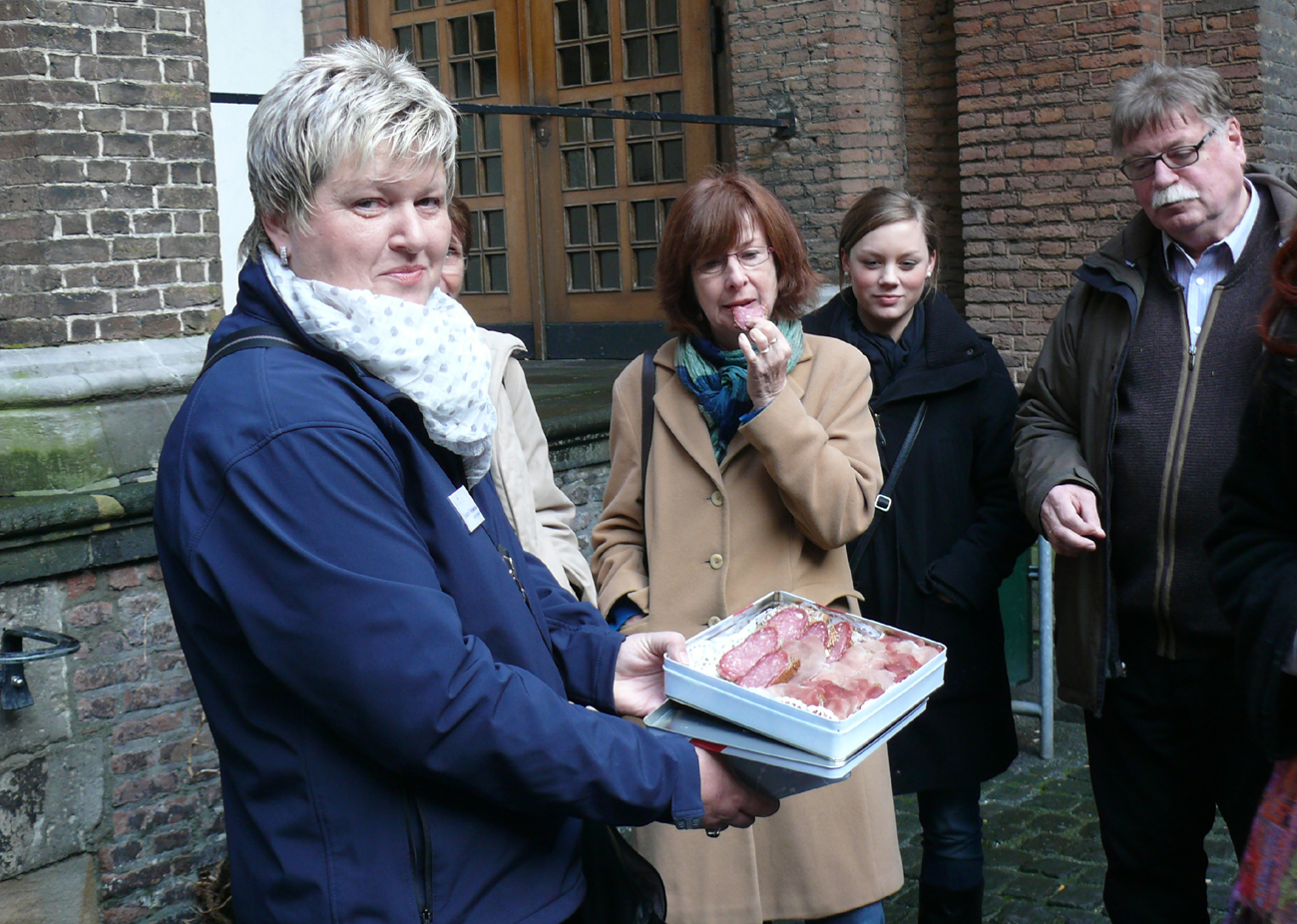 Person hält eine Schale mit mehreren Scheiben Wurst, umgeben von mehreren Personen vor einem Backsteingebäude.