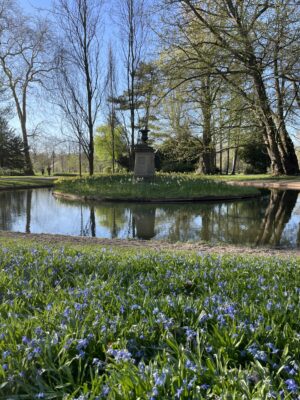 Parklandschaft mit kleinem Teich, Insel mit Denkmal und blühenden Blumen im Vordergrund unter Bäumen.