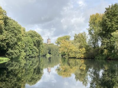 Fluss mit spiegelnder Wasseroberfläche, umgeben von Bäumen, im Hintergrund ein Kirchturm unter bewölktem Himmel.