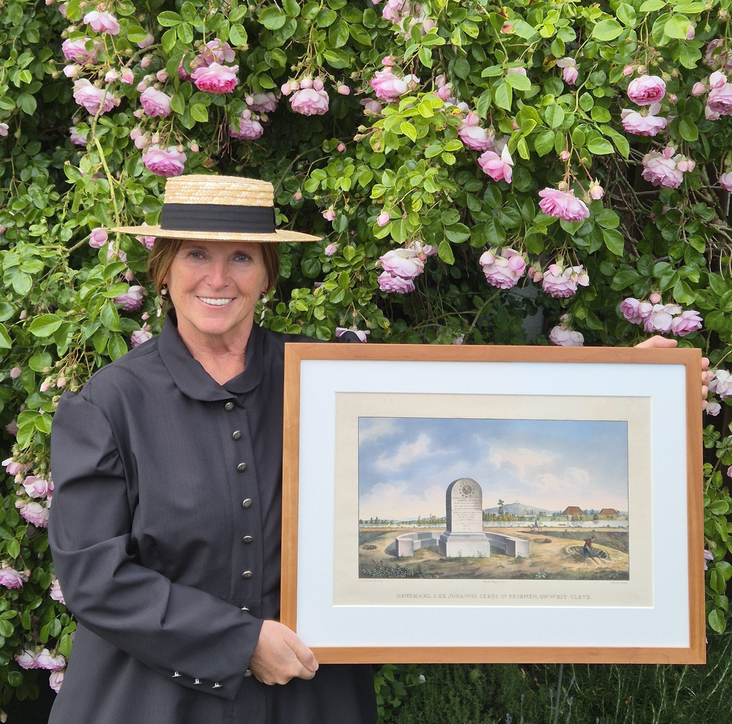 Stadtführerin Hildegard Liebeton in schwarzem Kleid und Strohhut hält gerahmtes Bild mit Grabstein vor blühendem Rosenstrauch.