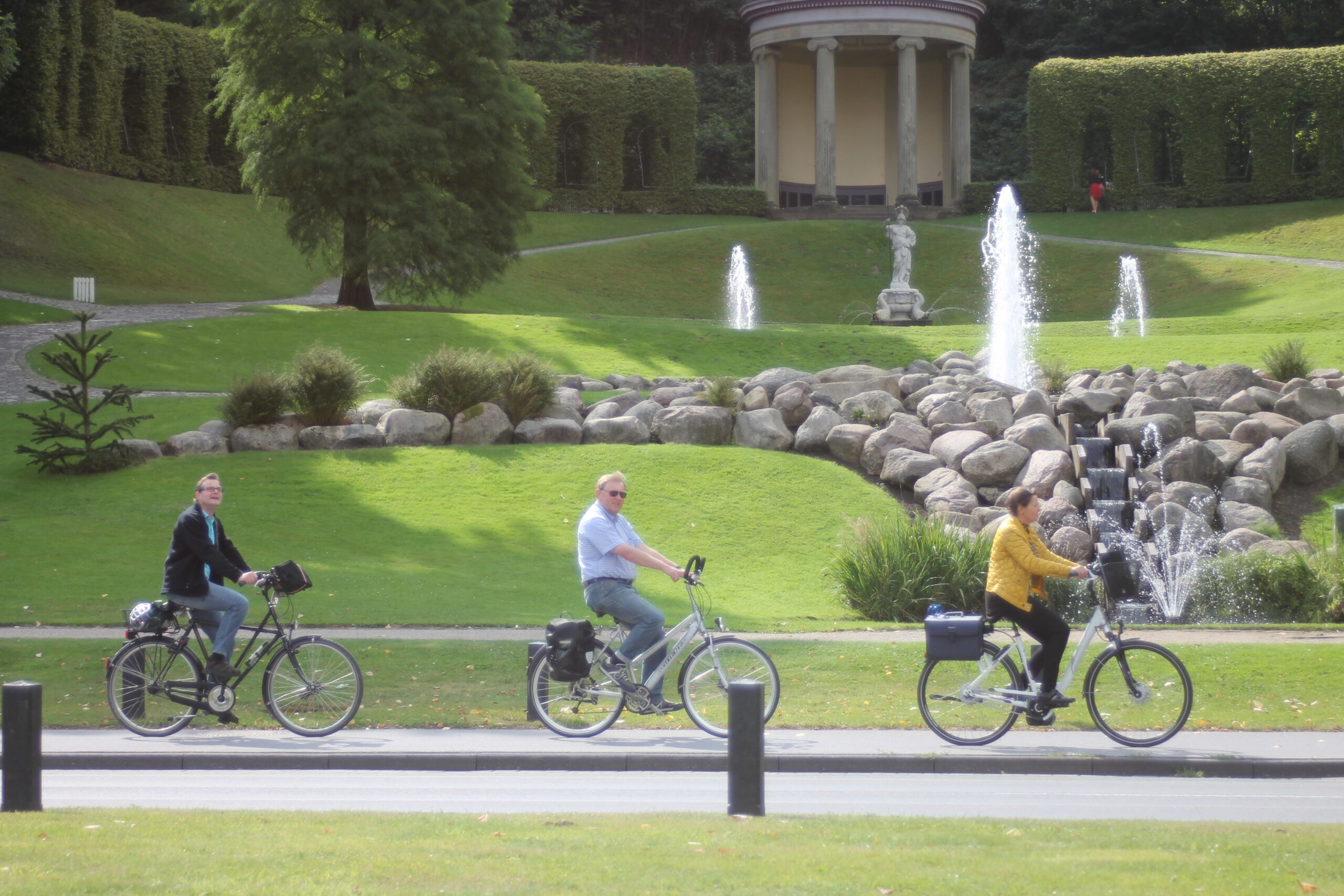 Drei Radfahrer fahren hintereinander auf einem Weg vor einem Garten mit Springbrunnen, Wasserfall und klassizistischem Pavillon.