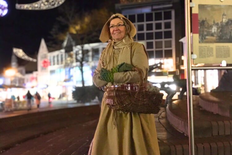Gästeführerin Birgit van den Boom in historischer Kleidung mit langem Mantel, Haube und Korb steht nachts auf gepflastertem Platz vor beleuchteten Gebäuden und Schwanenturm.