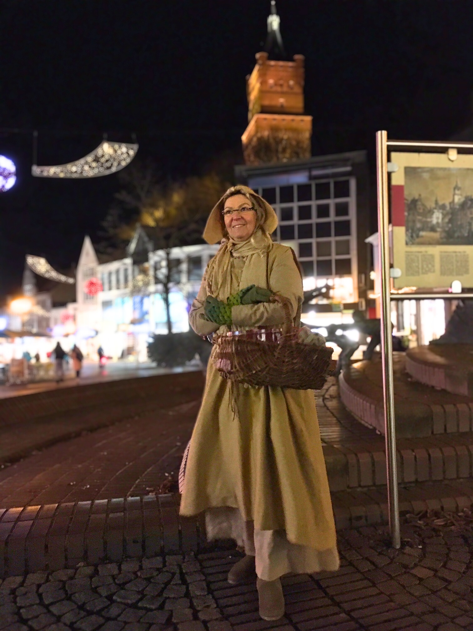 Gästeführerin Birgit van den Boom in historischer Kleidung mit langem Mantel, Haube und Korb steht nachts auf gepflastertem Platz vor beleuchteten Gebäuden und Schwanenturm.