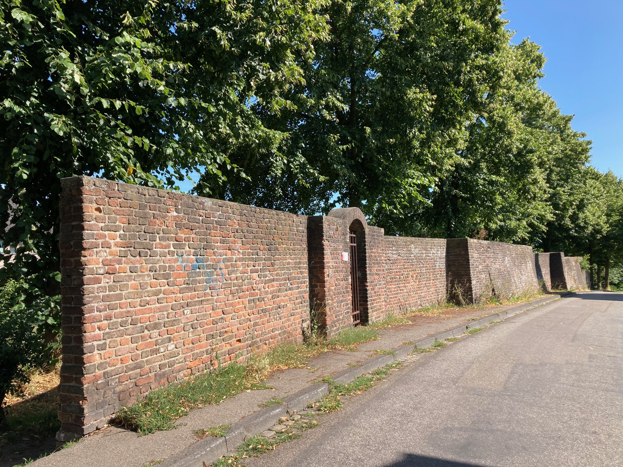 Lange Backsteinmauer mit einem kleinen Tor, umgeben von Bäumen und einem Gehweg daneben.