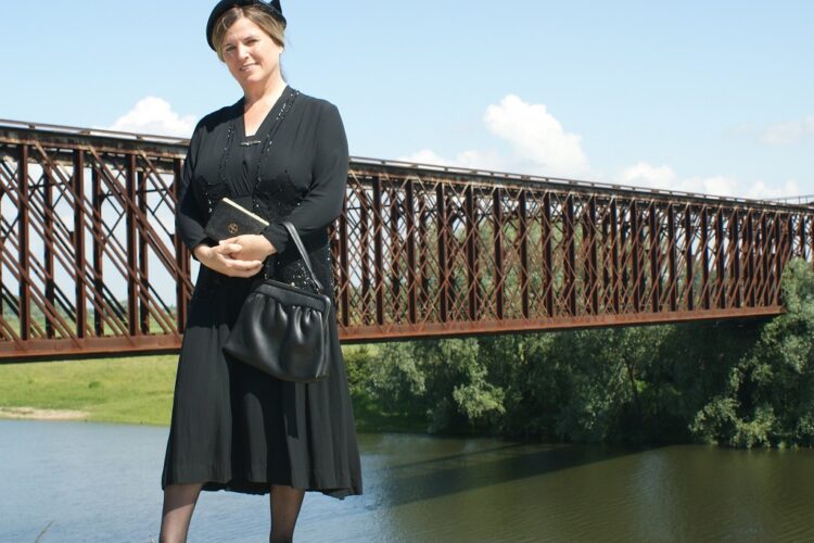 Frau in schwarzem Kleid, Hut und Schuhen steht vor einer Eisenbahnbrücke über einem Fluss.