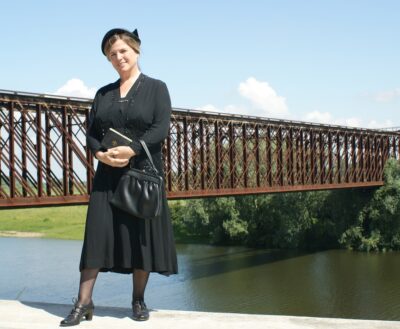 Frau in schwarzem Kleid, Hut und Schuhen steht vor einer Eisenbahnbrücke über einem Fluss.