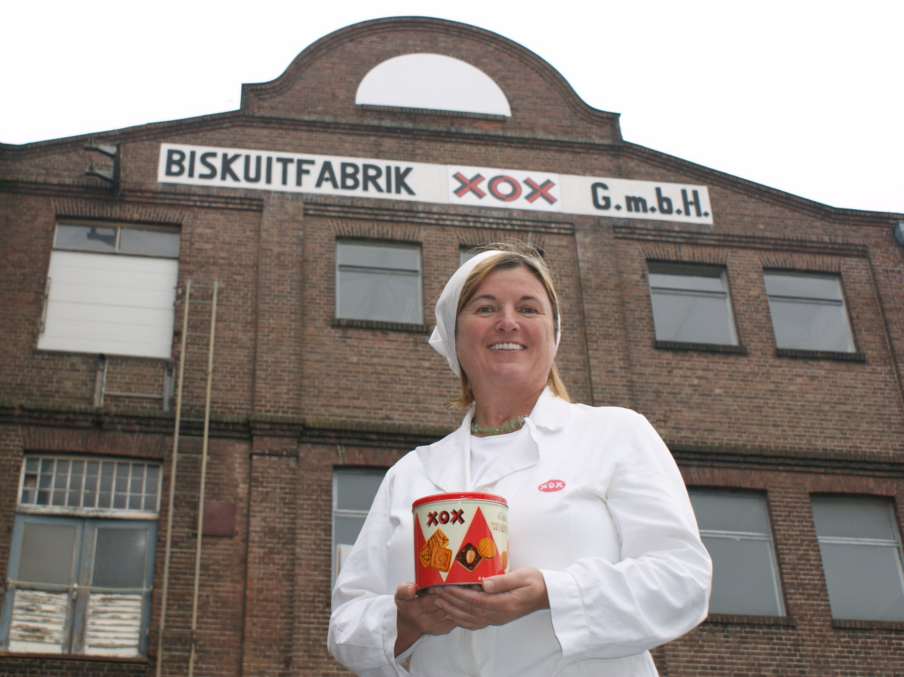 Stadtführerin Frau Liebeton in weißer Arbeitskleidung hält eine runde Blechdose mit der Aufschrift "XOX" vor einem Backsteingebäude mit dem Schriftzug "BISKUITFABRIK XOX G.m.b.H.".