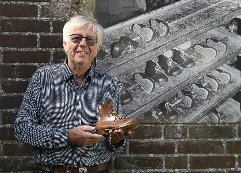 Gästeführer Norbert Leenders hält einen braunen Lederstiefel vor einer Backsteinwand mit einem schwarz-weißen Foto von mehreren Paar Schuhen auf einem Holzregal