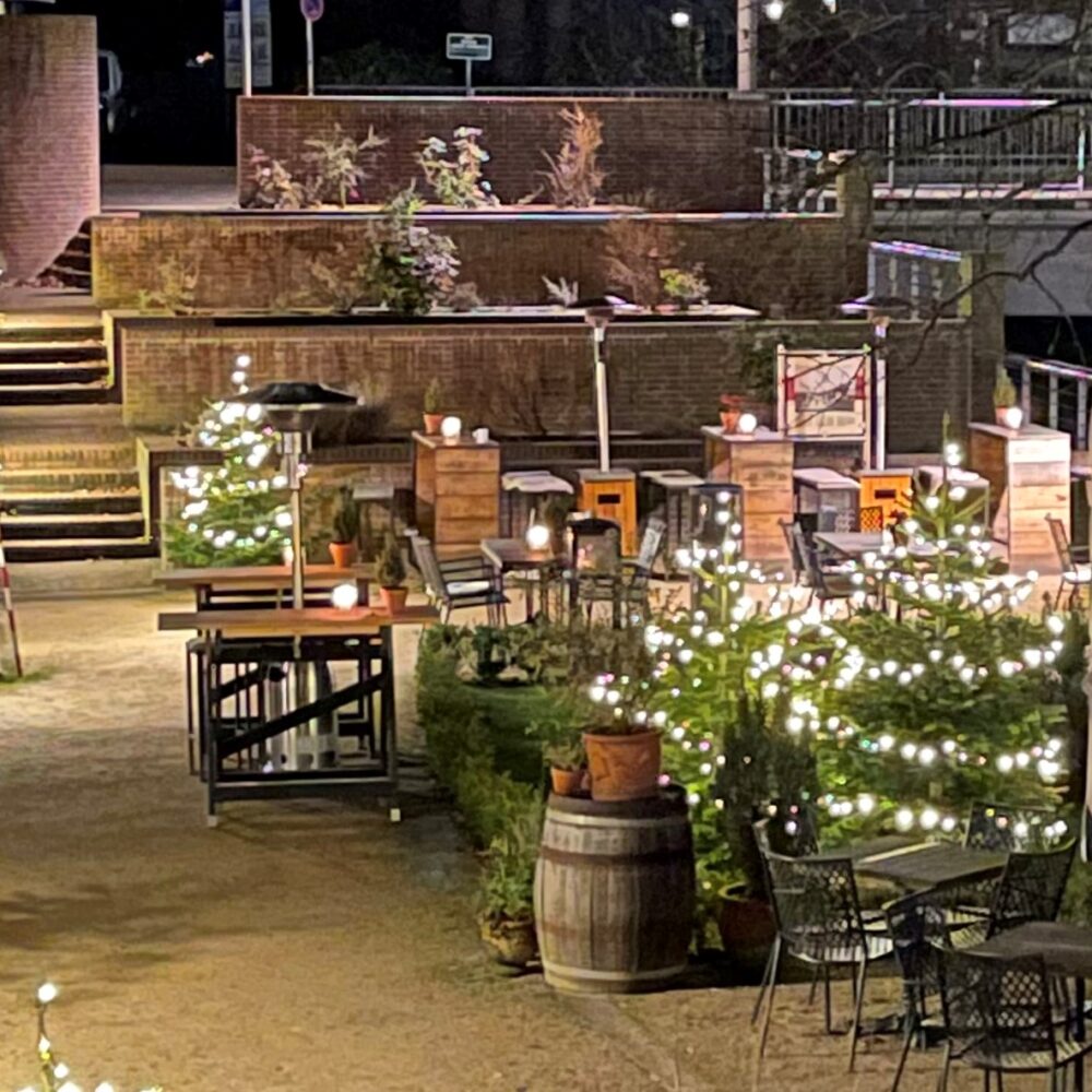 Beleuchtete Tische und Stühle in einer Außenwirtschaft mit kleinen Tannenbäumen und Lichterketten bei Nacht.