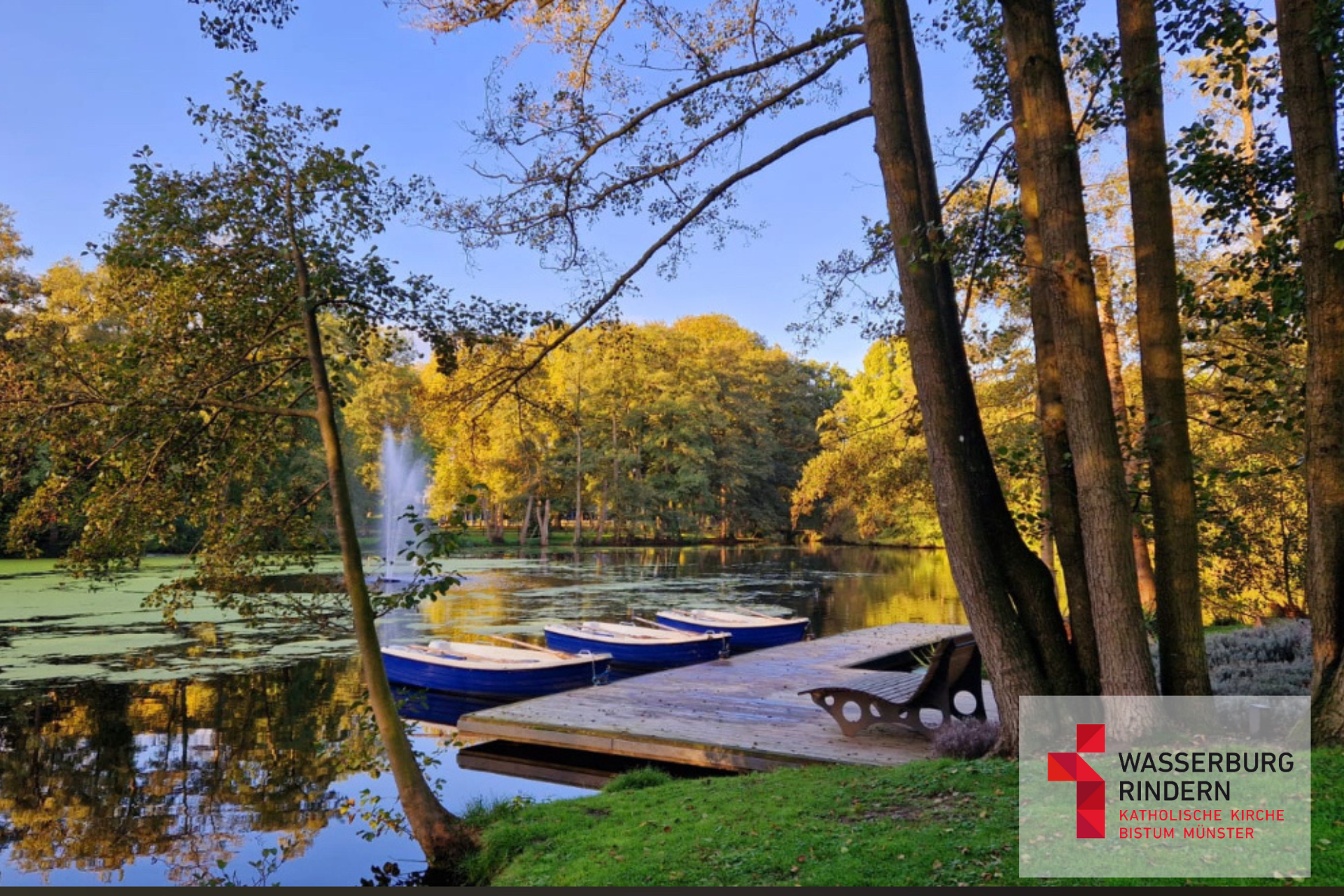 Anlegesteg der Wasserburg Rindern mit drei Booten und Liegestühlen. Um den See herum stehen Bäume und in der Mitte des Sees erhebt sich eine Fontäne aus dem Wasser.