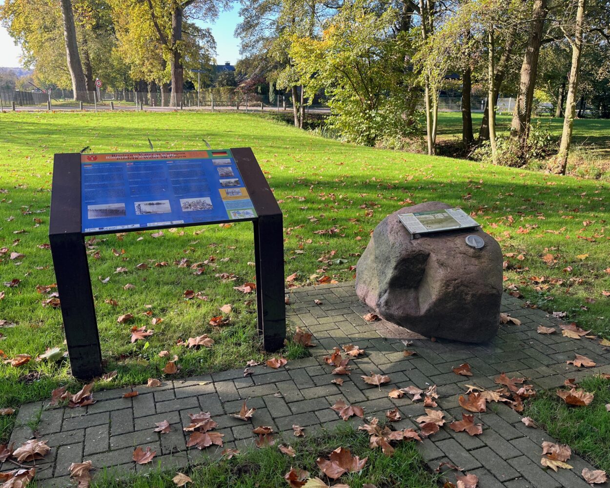 Infotafel und Findling im Grünen, im Hintergrund Bäume