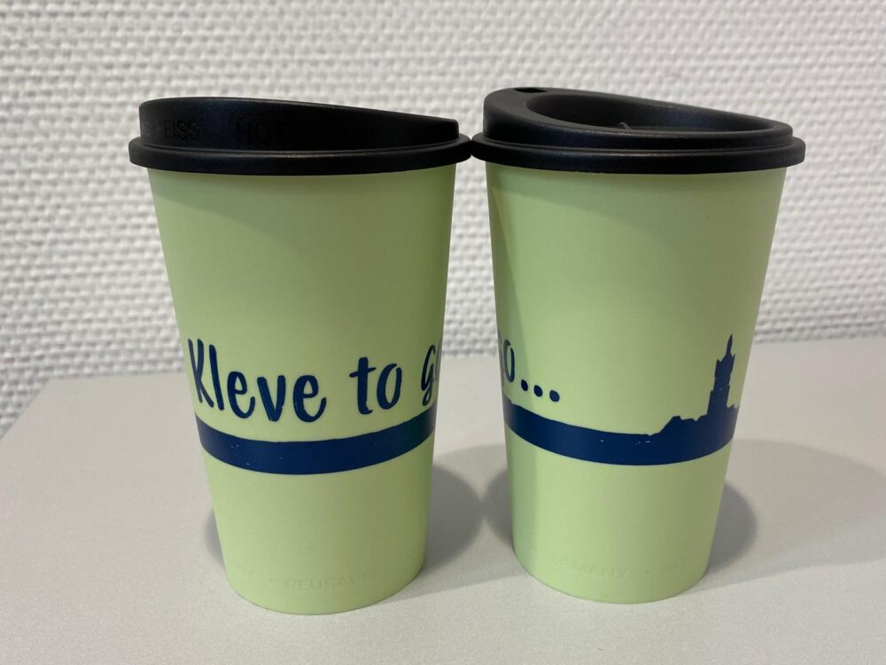 Zwei hellgrüne Mehrwegbecher mit schwarzen Deckeln, bedruckt mit dunkelblauer Schrift "Kleve to go" und einer stilisierten Stadtsilhouette der Klever Schwanenburg.
