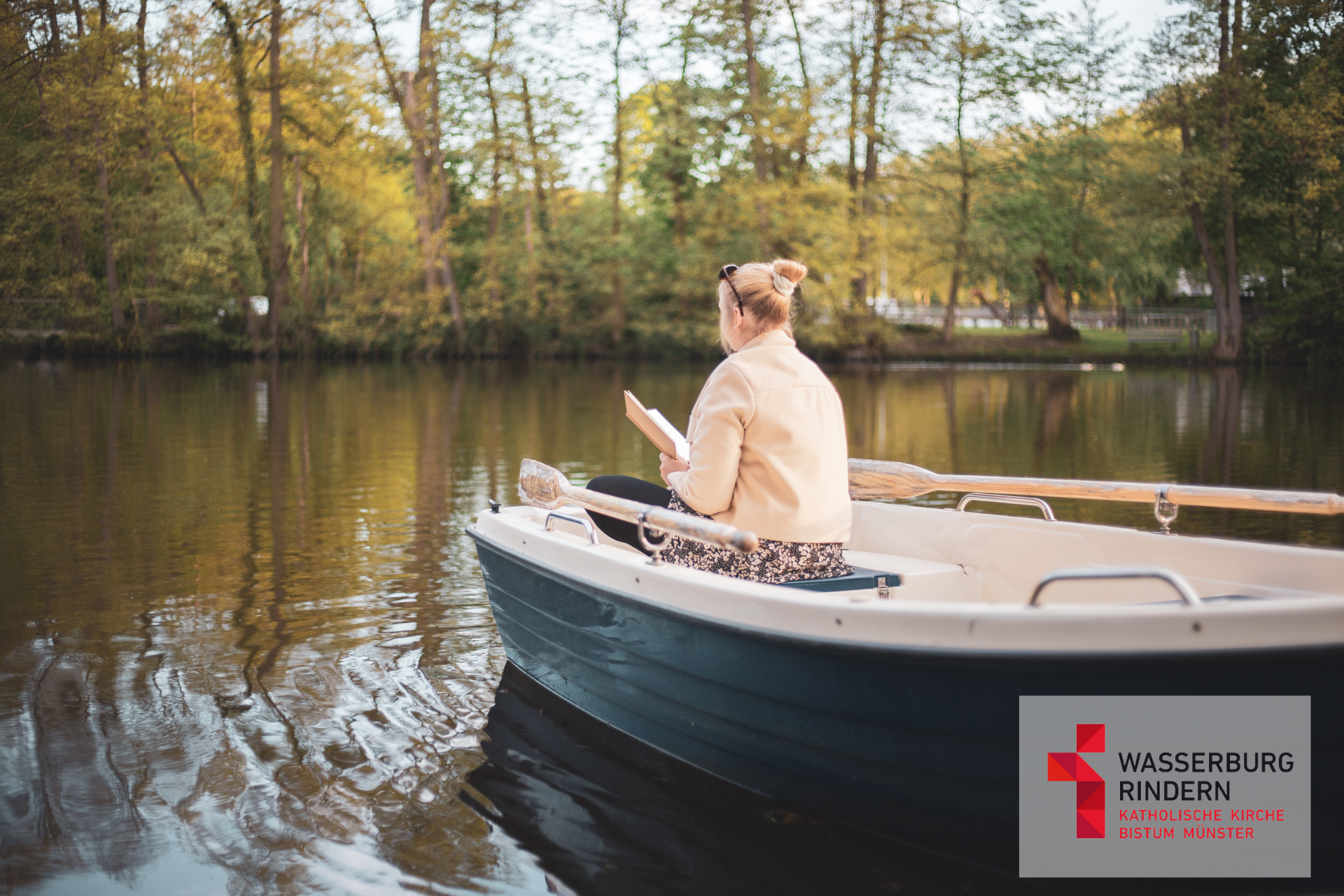 Frau liest ein Buch in einem Ruderboot auf einem See der von Bäumen gesäumt ist.