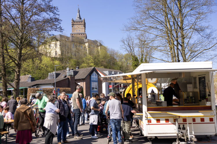 Menschen stehen an einem Imbisswagen in einer belebten Straße mit Kirschblütenbäumen und der Schwanenburg im Hintergrund
