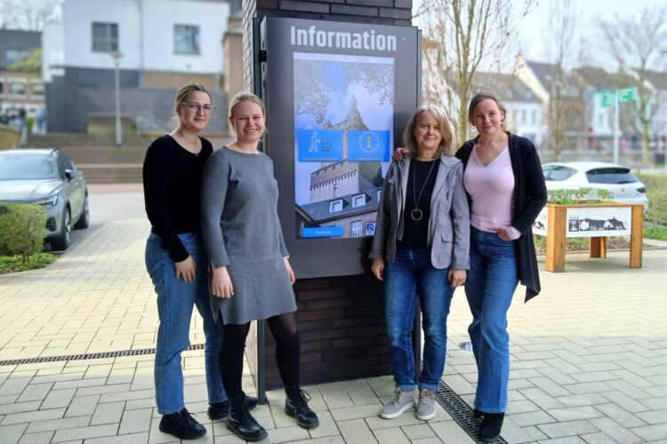 Vier Mitarbeiterinnen aus dem Team Tourismus stehen vor der Infostele vor der Tourist Information Kleve