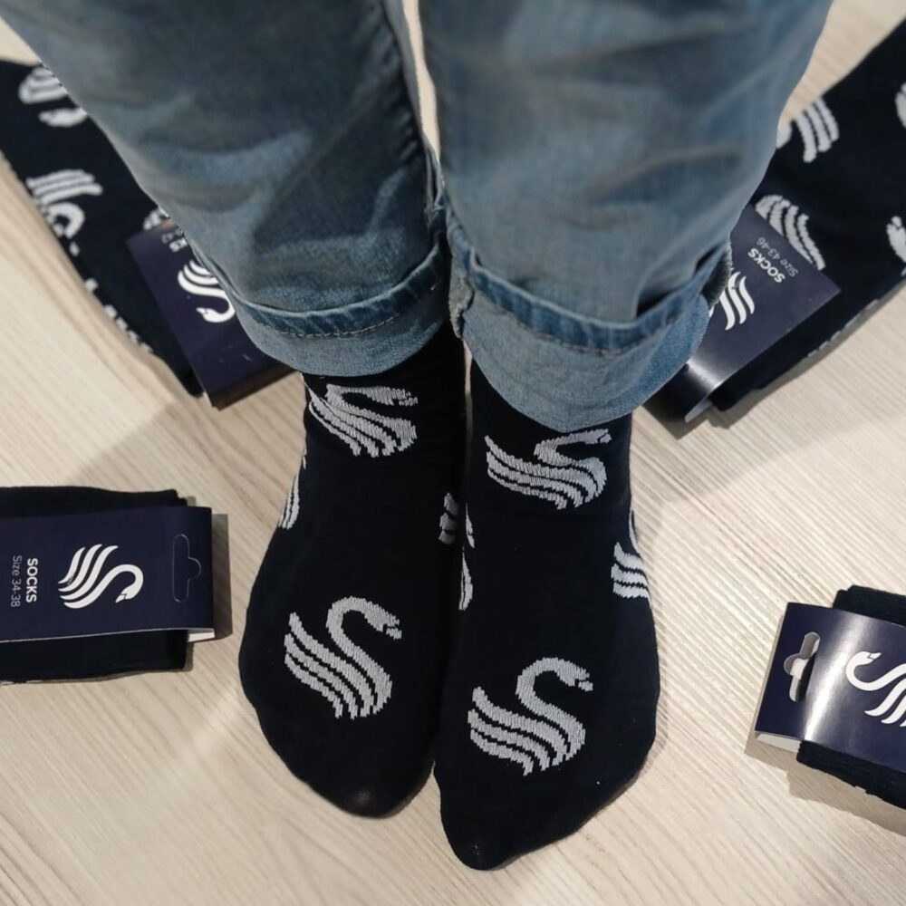 Person trägt dunkelblaue Socken mit weißem geschwungenem Muster, umgeben von mehreren verpackten Socken mit gleichem Design auf hellem Boden