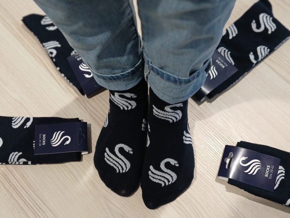 Person trägt dunkelblaue Socken mit weißem geschwungenem Muster, umgeben von mehreren verpackten Socken mit gleichem Design auf hellem Boden