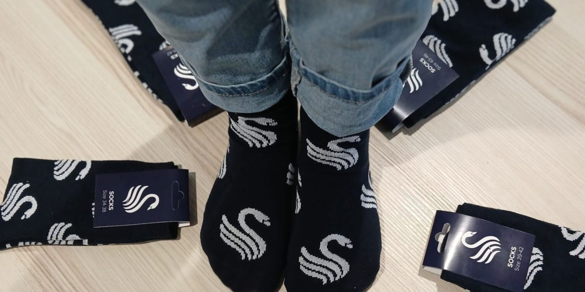 Person trägt dunkelblaue Socken mit weißem geschwungenem Muster, umgeben von mehreren verpackten Socken mit gleichem Design auf hellem Boden