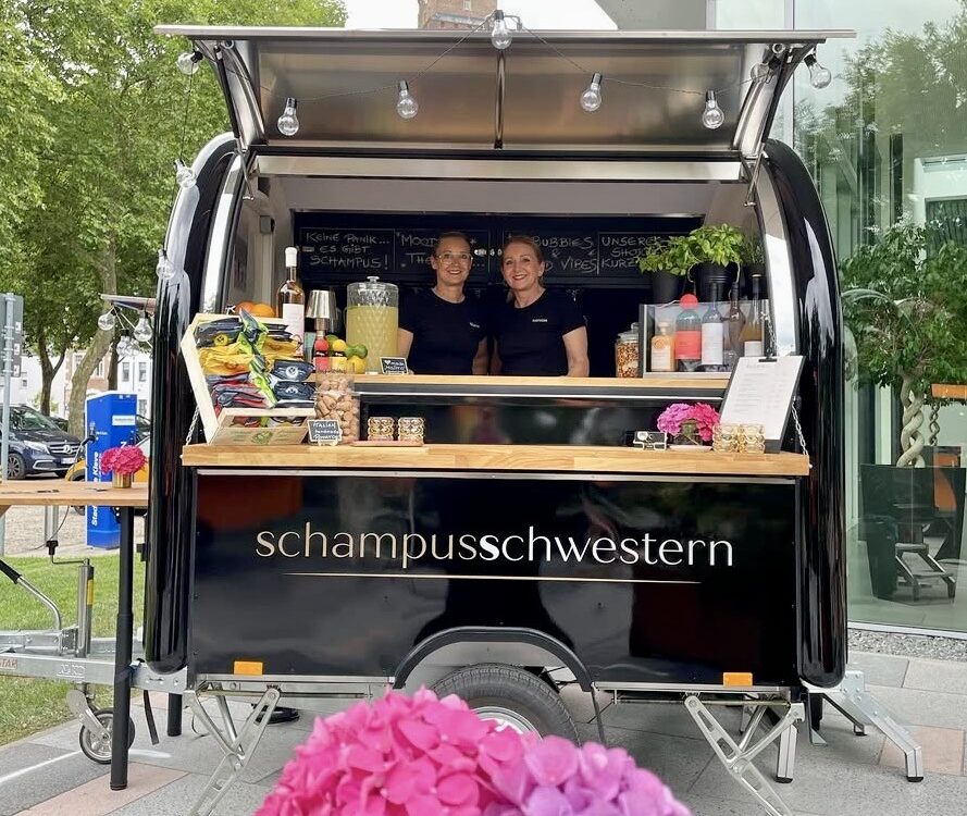 Mobiler schwarzer Verkaufsanhänger mit der Aufschrift »Schampusschwestern« und zwei Personen hinter dem Tresen, davor ein Tisch mit rosa und lila Hortensien in einer Vase