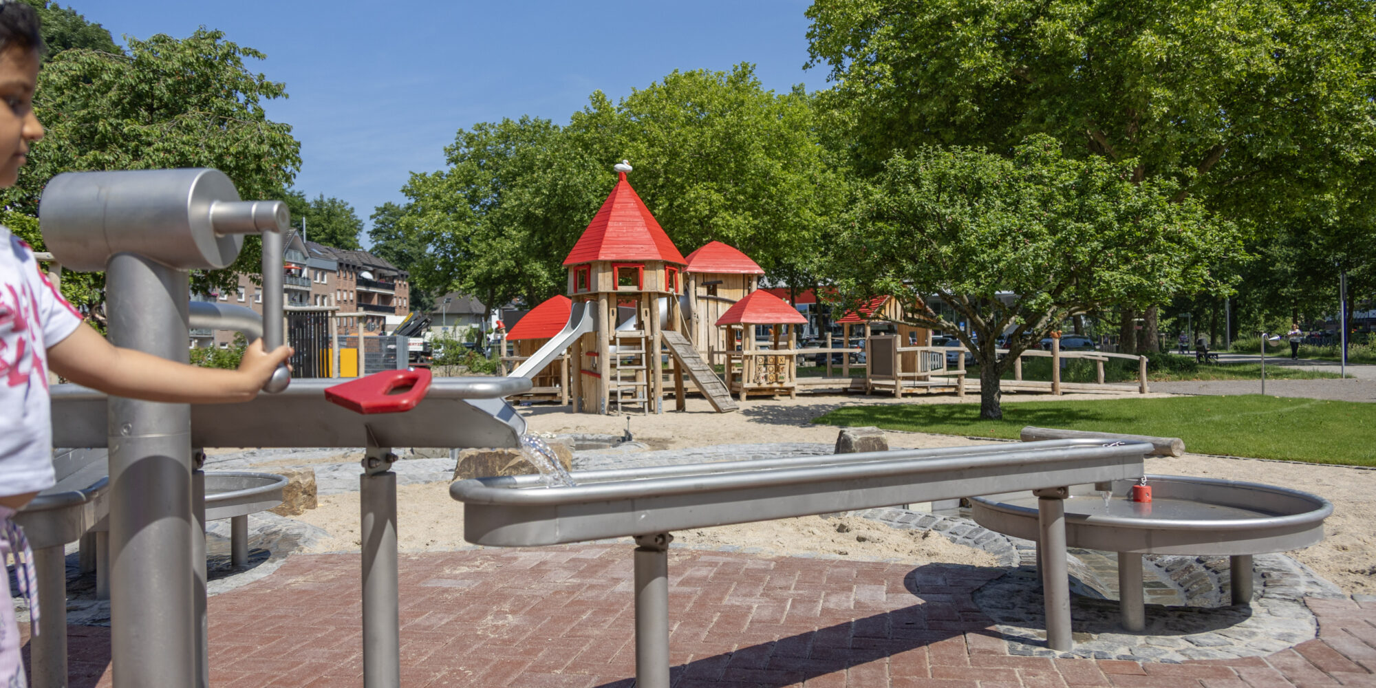 Kind bedient Wasserspielanlage mit rotem Griff auf Spielplatz mit Holzturm und grünen Bäumen im Hintergrund