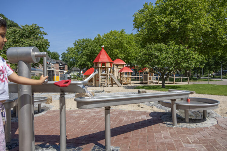 Kind bedient Wasserspielanlage mit rotem Griff auf Spielplatz mit Holzturm und grünen Bäumen im Hintergrund