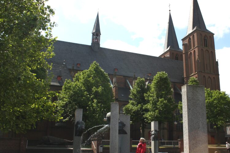 Stiftskirche mit zwei hohen Türmen und kleinem Turm in der Mitte, davor mehrere Steinsäulen die den Narrenbrunnen bilden und eine weibliche Person in rotem Mantel