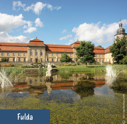 Barockes Schloss mit rotem Dach und Turm spiegelt sich in einem Teich mit Skulptur und Pflanzen