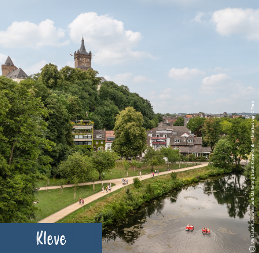 Flussufer mit Spazierweg, Bäumen und Menschen, im Hintergrund Häuser und ein hoher Turm auf einem bewaldeten Hügel