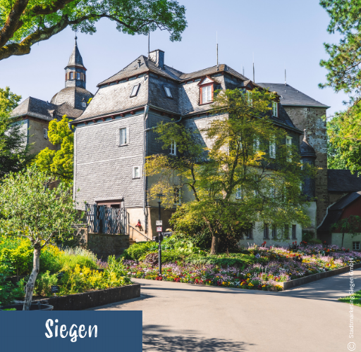 Mehrstöckiges historisches Gebäude mit Schieferfassade und Türmen umgeben von Bäumen und Blumenbeeten an sonnigem Tag