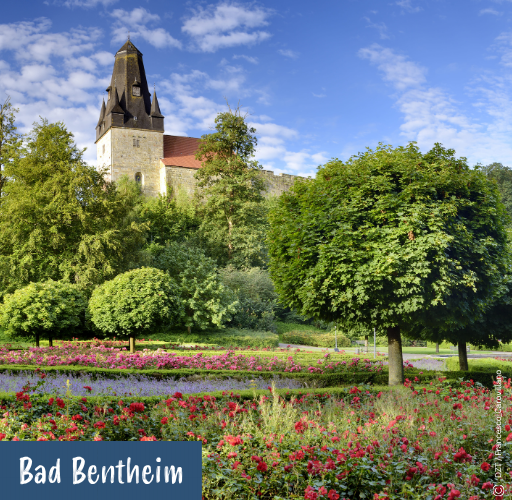 Kirchturm hinter Bäumen mit Blumenbeeten im Vordergrund und blauem Himmel