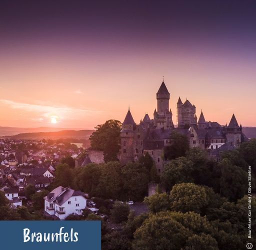 Burg auf einem bewaldeten Hügel mit Häusern im Vordergrund bei Sonnenuntergang