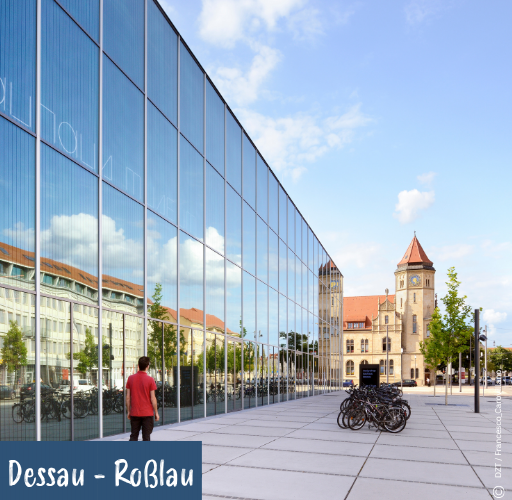 Moderne Glasfassade spiegelt historische Gebäude und Himmel, davor stehen Fahrräder und eine Person in rotem T-Shirt