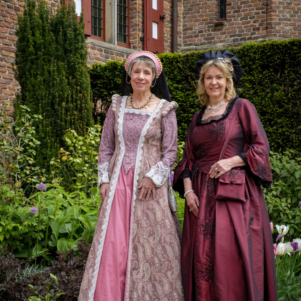 Zwei Frauen in historischen Kleidern, Maria von Nassau links und Luise Henriette von Oranien rechts, stehen nebeneinander auf einem Grasstreifen vor einer Hecke, links eine Frau in einem hellen, gemusterten Kleid mit Kopfschmuck, rechts eine Frau in einem dunklen, glänzenden Kleid mit Haube.