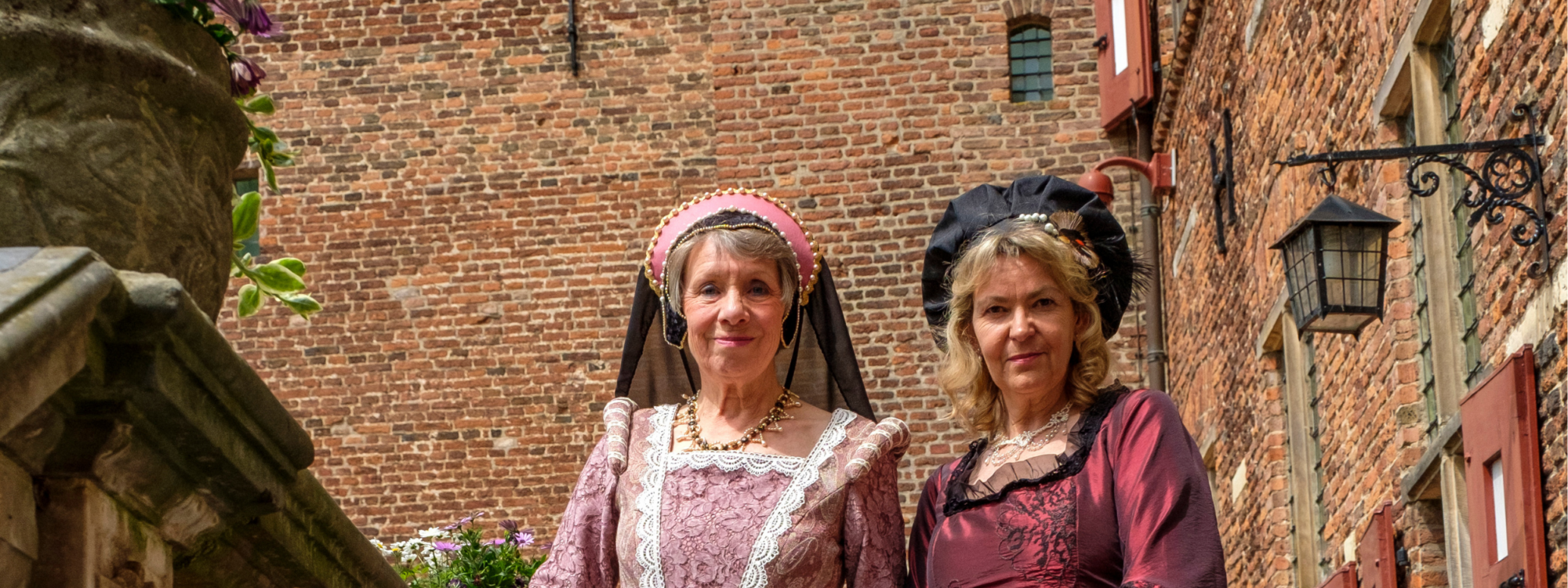 Zwei Frauen in historischen Kleidern, Maria von Nassau links und Luise Henriette von Oranien rechts, stehen nebeneinander auf einem gepflasterten Weg vor einer Backsteinmauer mit roten Fensterläden, links eine Frau in einem hellen, gemusterten Kleid mit Kopfschmuck, rechts eine Frau in einem dunklen, glänzenden Kleid mit Haube.