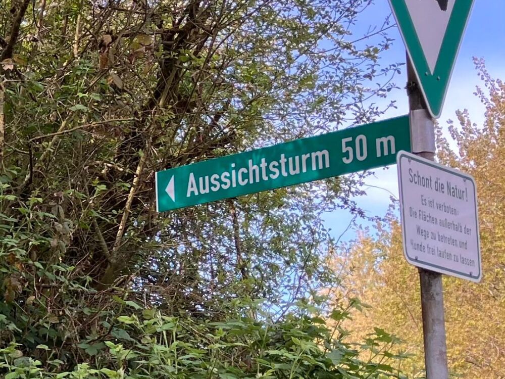 Wegweiser mit der Aufschrift 'Aussichtsturm 50 m' und Naturschutzgebiet-Schild an einem Pfahl vor Büschen und Bäumen