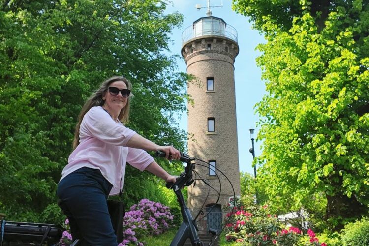 Frau mit Sonnenbrille auf schwarzem Fahrrad vor einem runden Steinturm und grünen Bäumen