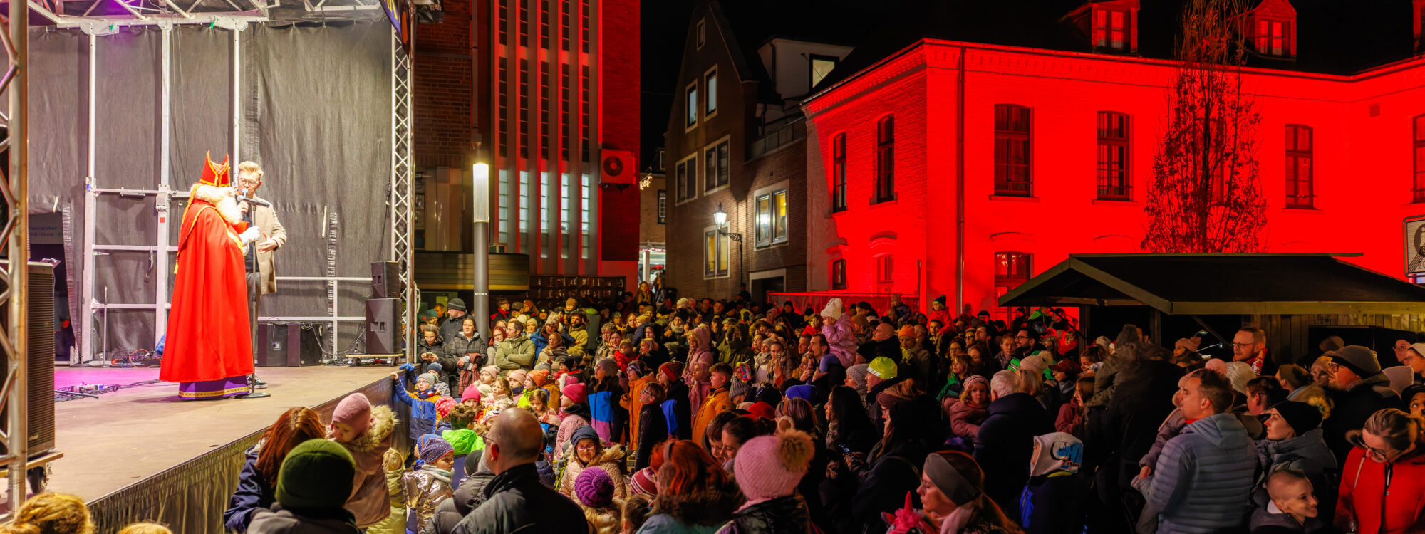 Menschenmenge vor Bühne mit zwei verkleideten Personen, eine als Nikolaus, in einem abendlichen Stadtplatz mit rot beleuchtetem Gebäude im Hintergrund