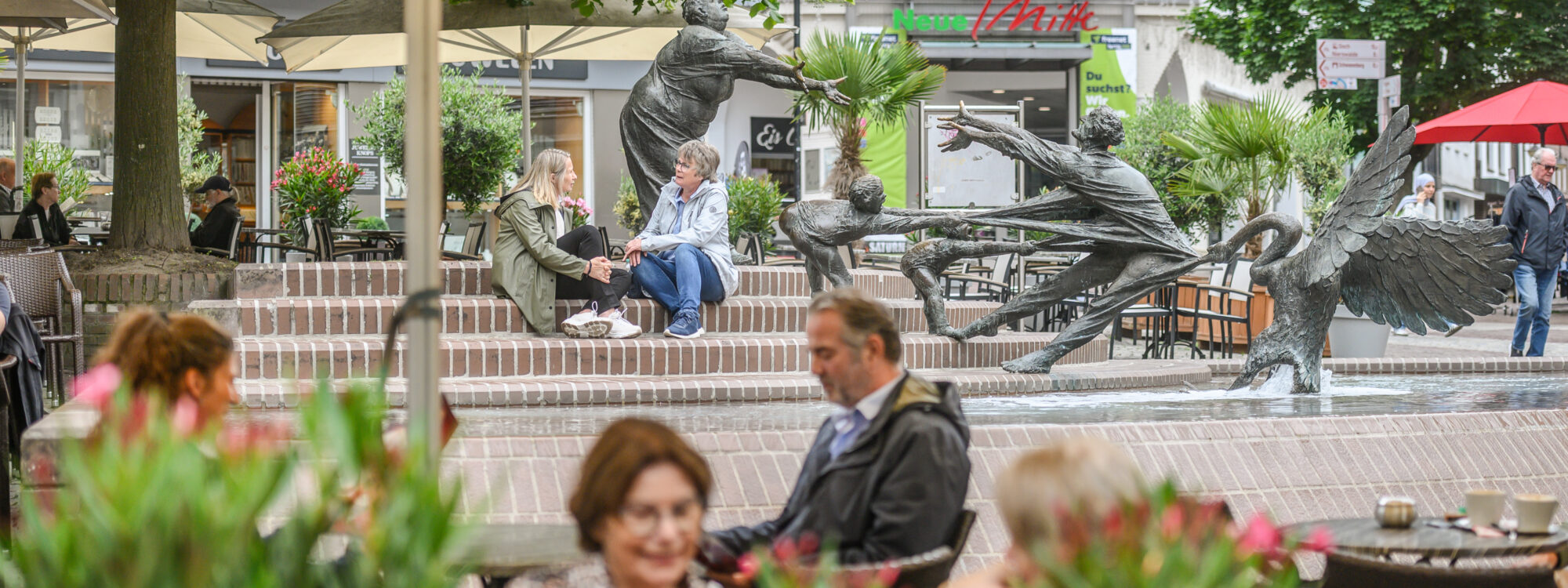 Brunnen mit mehreren dynamischen Figuren in einer belebten Innenstadt mit Sitzgelegenheiten und Pflanzen im Vordergrund