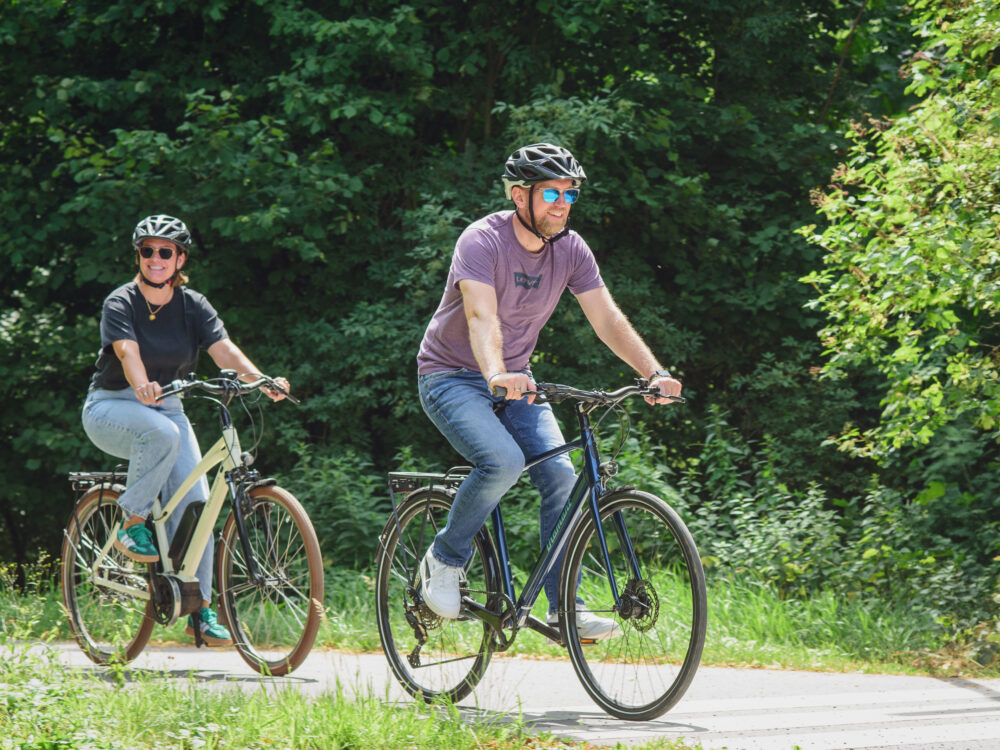 Zwei Personen mit Fahrradhelmen fahren auf einem Weg vor grünem Laubwald, vorne ein Mann in lila T-Shirt und Jeans, dahinter eine Frau in schwarzem T-Shirt und Jeans.