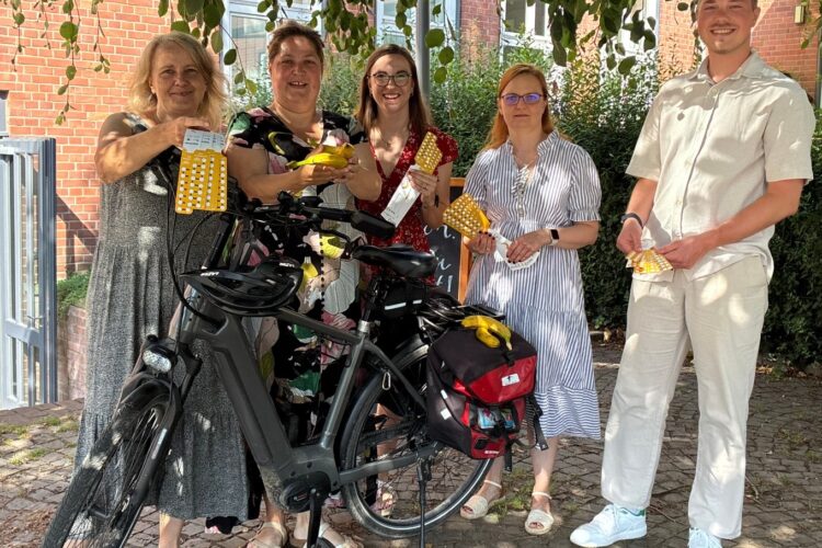 Fünf Personen stehen im Schatten eines Baumes hinter einem Fahrrad und halten gelbe Bänder in die Kamera