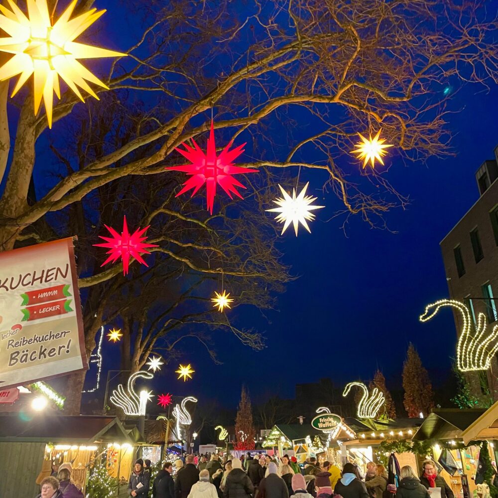 Mehrere leuchtende Weihnachtssterne in Gelb, Rot und Weiß hängen an kahlen Ästen über dem belebten Klever Weihnachtsmarkt mit Buden und Menschen bei Nacht