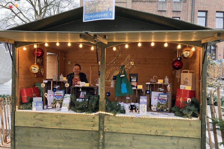 Holzstand mit Weihnachtsdekoration, Geschenken und einer Person hinter dem Tresen unter einem Schild mit der Aufschrift 'Vereinsbude 1'