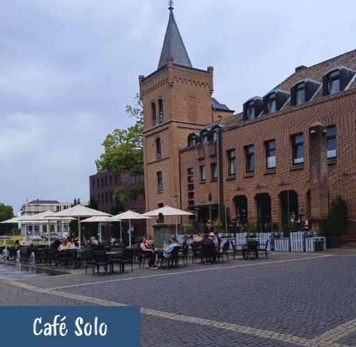 Außenbereich eines Cafés mit Tischen, Stühlen und Sonnenschirmen vor einem mehrstöckigen Backsteingebäude mit Turm