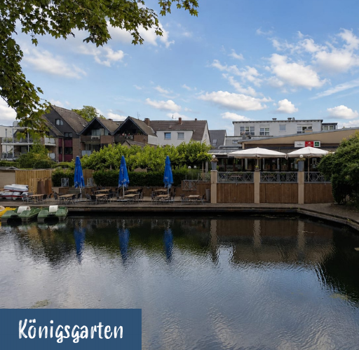 Blick auf einen Fluss mit Sitzbereich und blauen Sonnenschirmen vor Wohnhäusern unter bewölktem Himmel