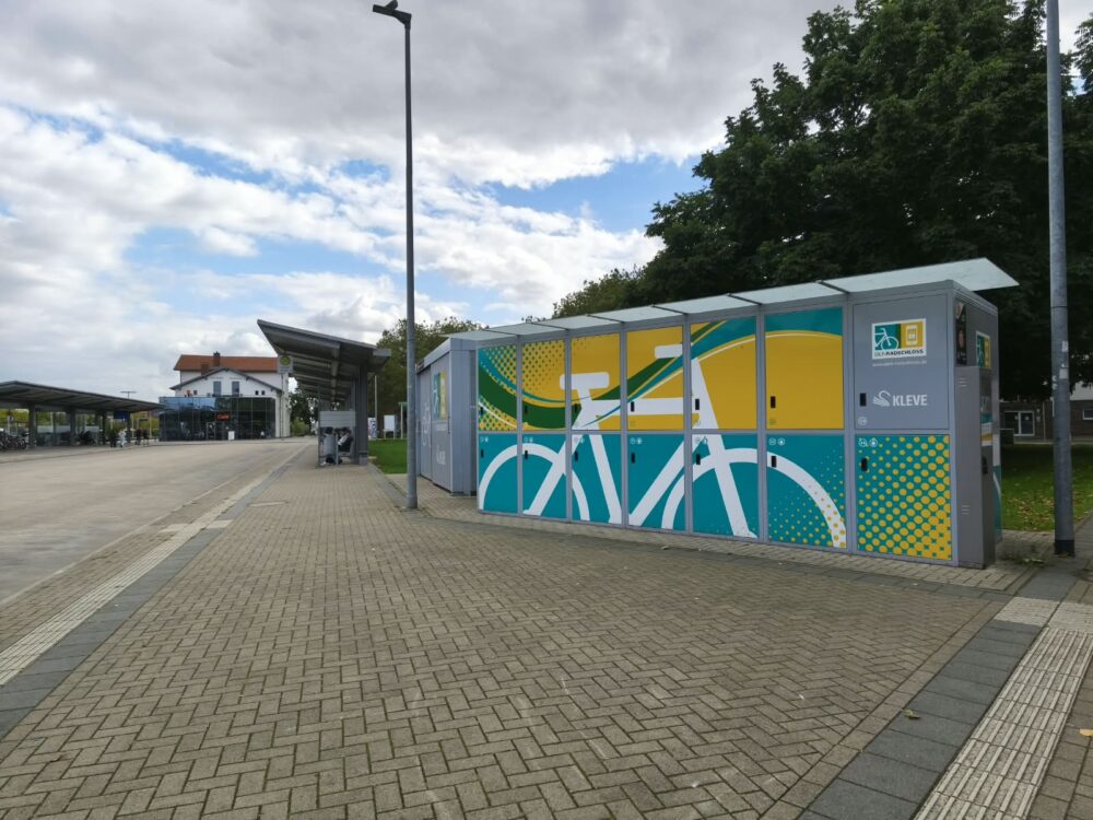 Mehrere Fahrradboxen mit farbiger Fahrradgrafik stehen auf einem gepflasterten Platz neben einem Bahnhof und Bäumen