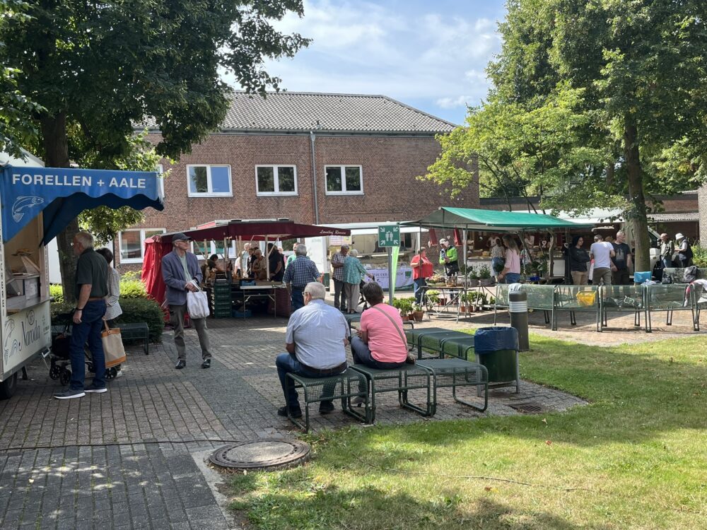 Markt mit Ständen und Menschen auf gepflastertem Platz vor Backsteingebäude, zwei Personen sitzen auf Bank