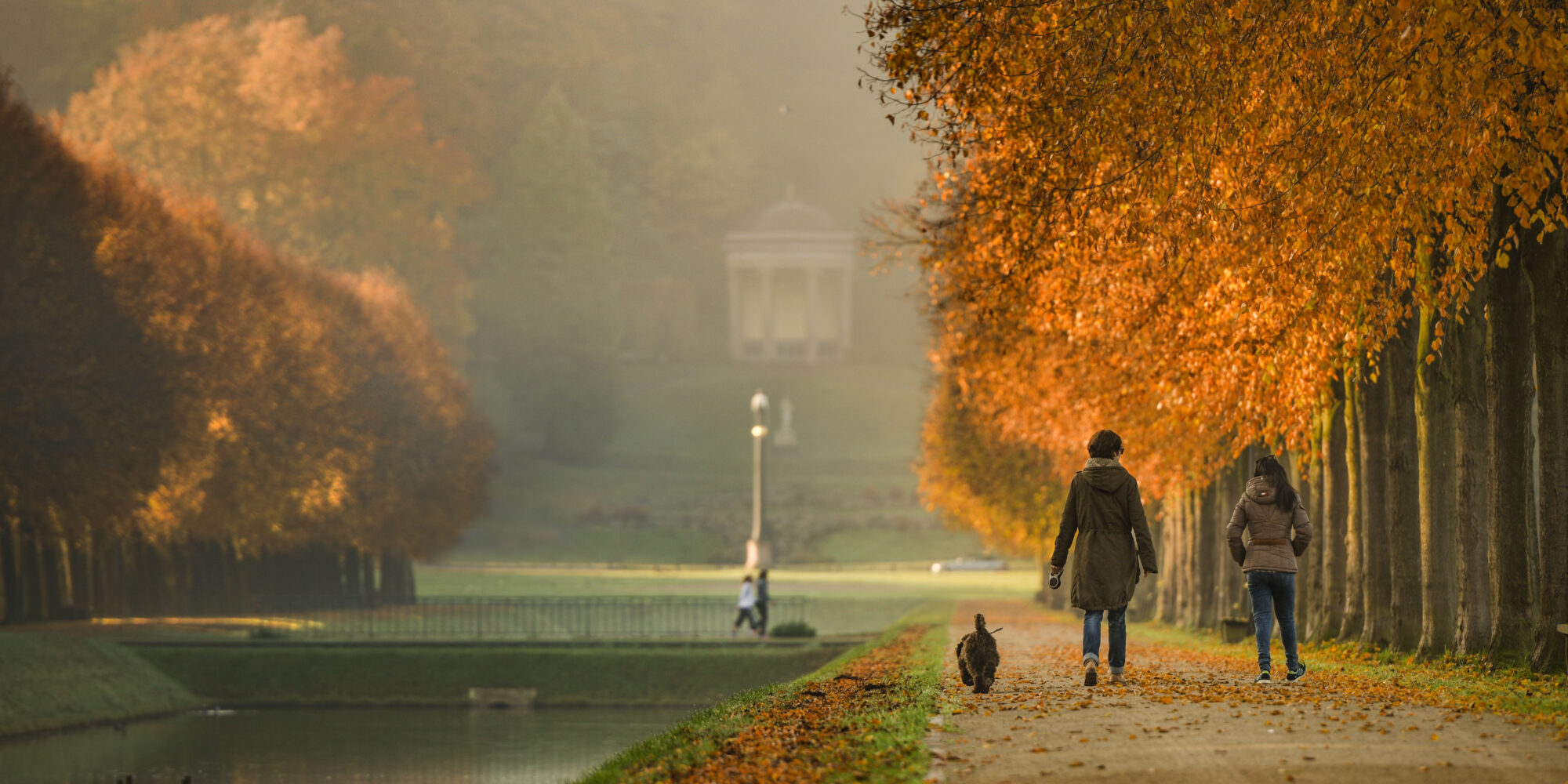 Zwei Frauen und ein Hund laufen durch die barocken Gartenanlagen. Die Bäume sind bunt gefärbt.